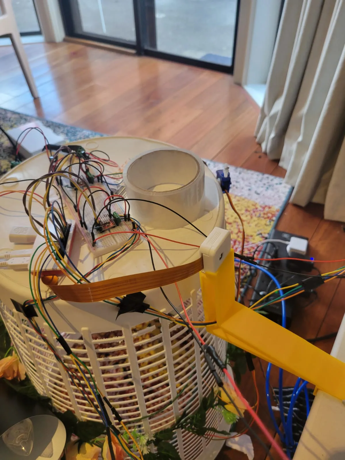 A breadboard covered in jumper wires sitting on top of an upturned laundry basket, with a camera module mounted on a yellow 3D printed arm.