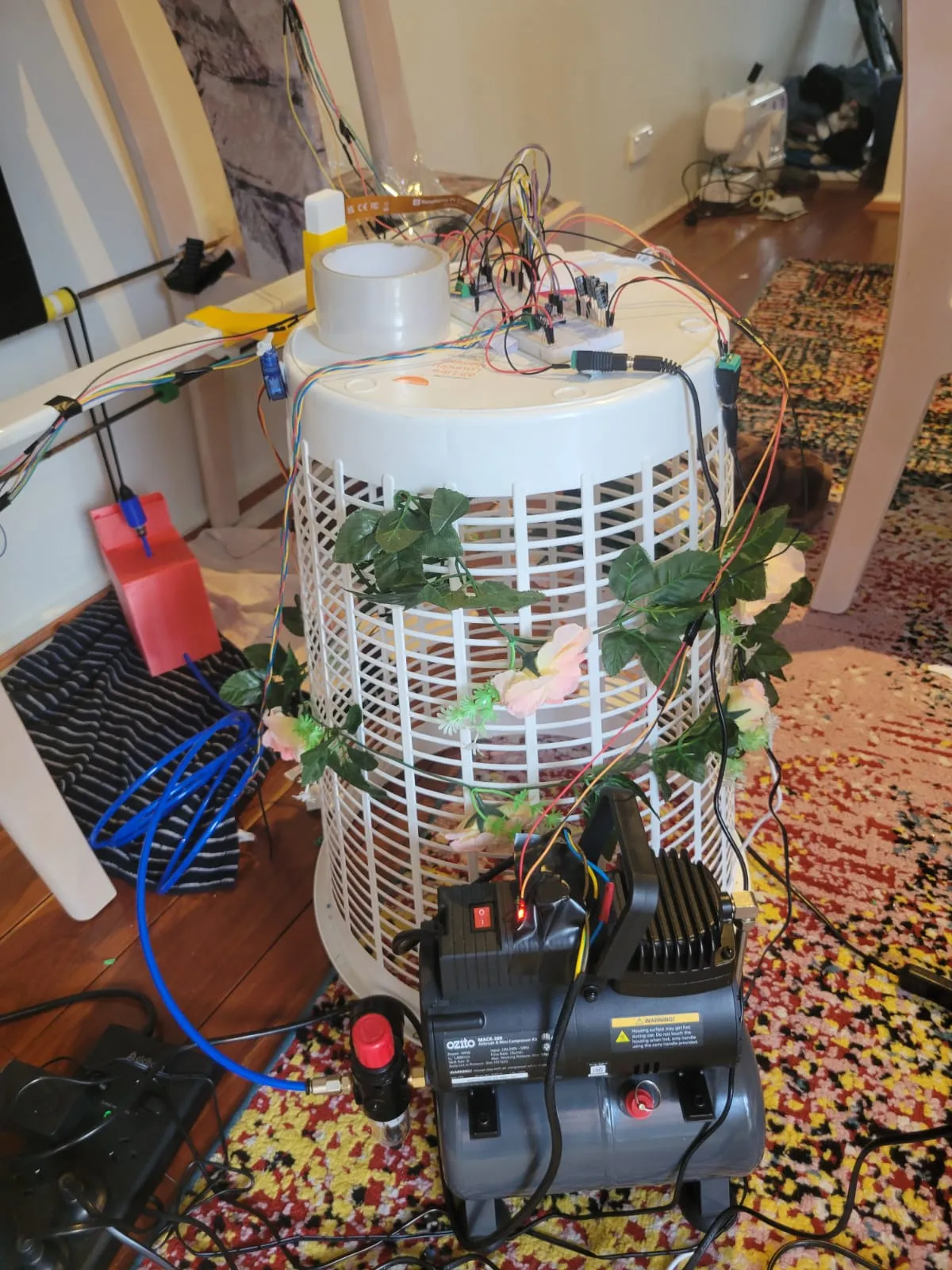 An Ozito airbrush compressor on the floor in front of an upturned laundry basket, connected by a blue air hose, with fake flowers threaded through the basket.