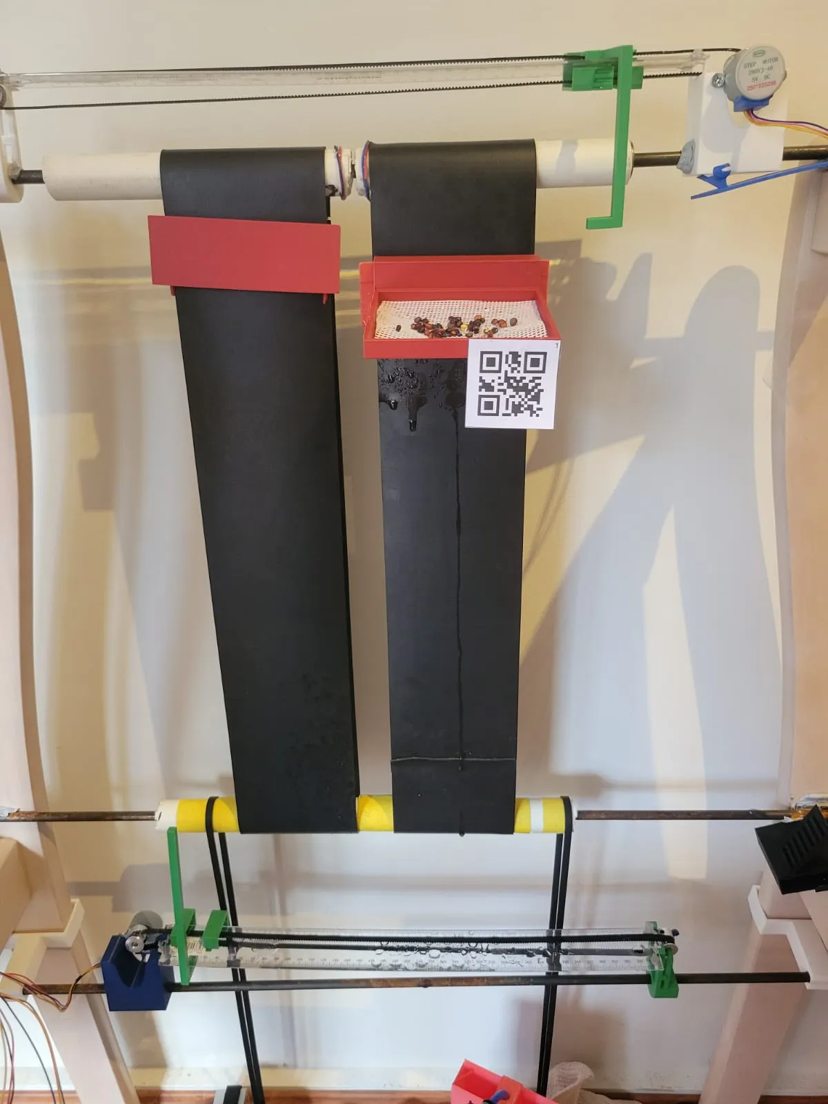 Two black rubber conveyor belts running vertically between white PVC rollers, with a red 3D printed tray of seeds hanging off one of them and a QR code taped beside it.