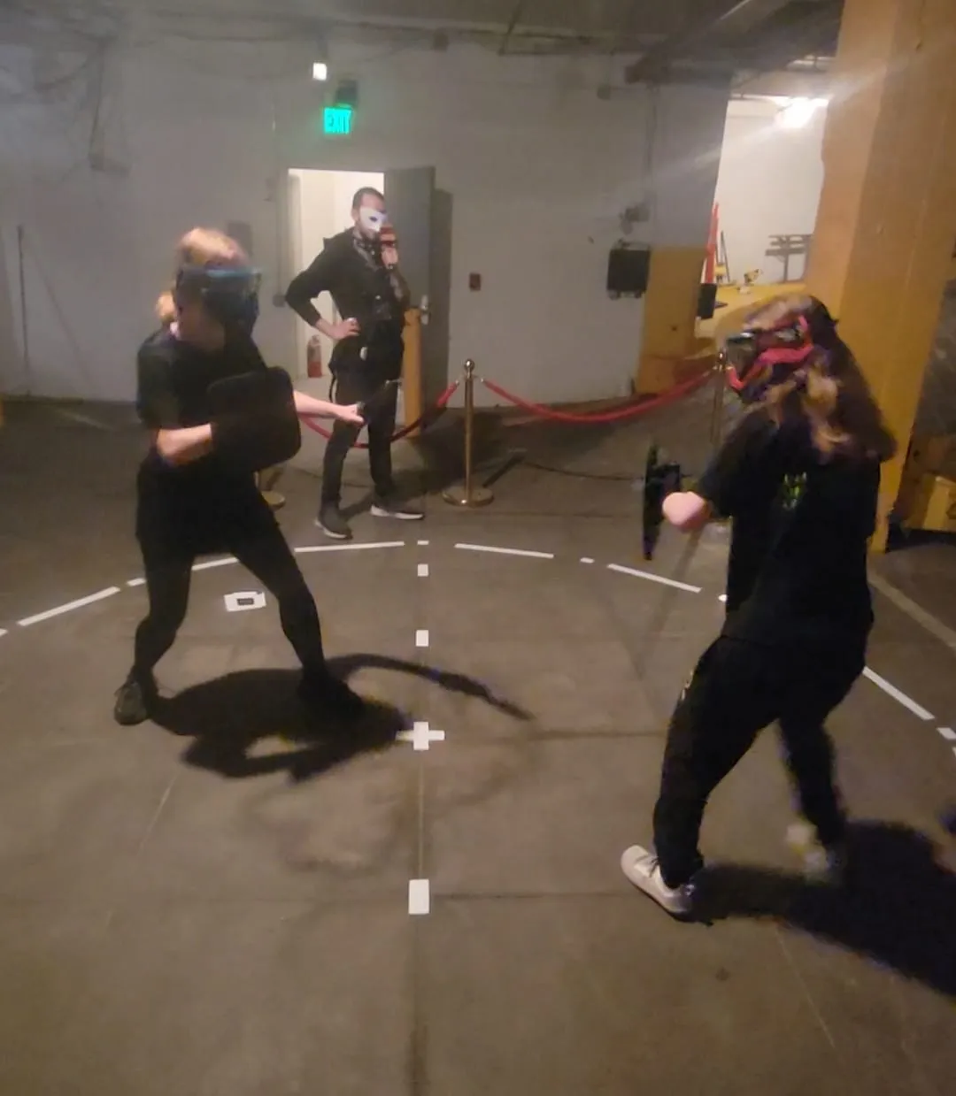 Two people in protective masks and all black circle each other with training knives inside a taped ring on a concrete floor, while a masked onlooker films from the edge.